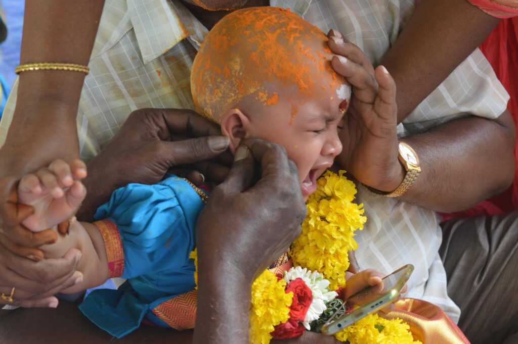 A baby in a traditional Indian ear-piercing ceremony, surrounded by family hands, flowers, and rituals.