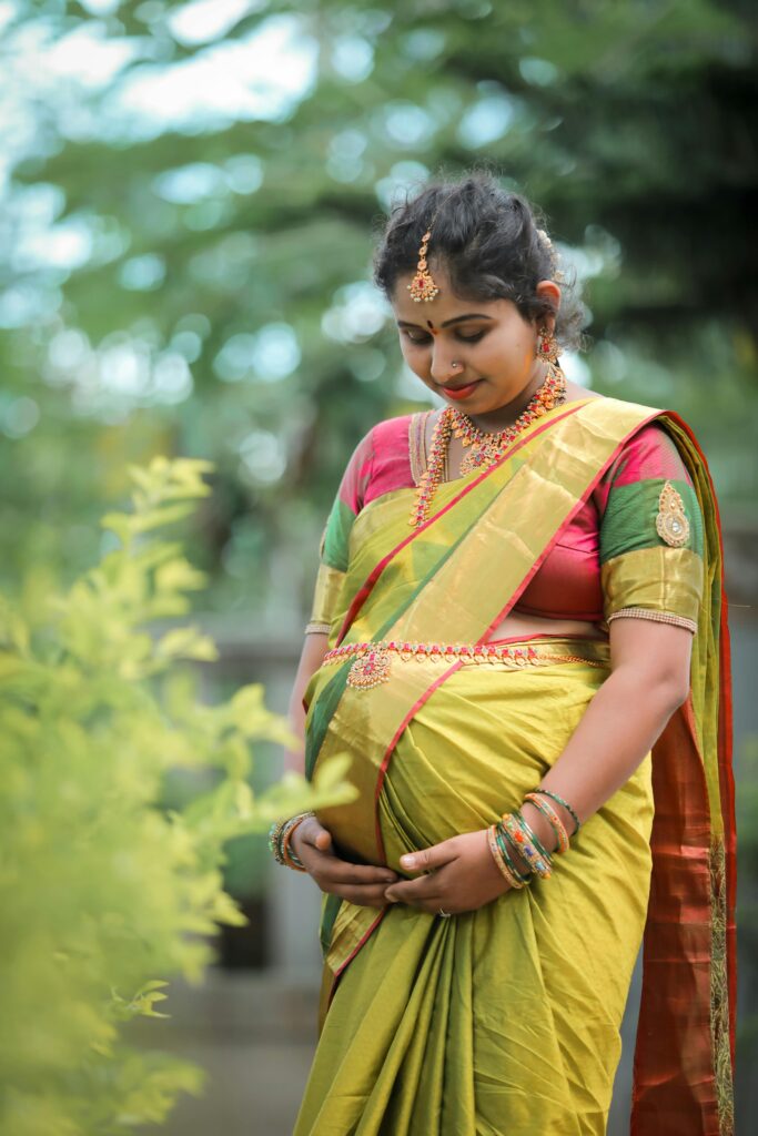 A pregnant woman dressed in a vibrant saree, smiling gracefully amid lush greenery.