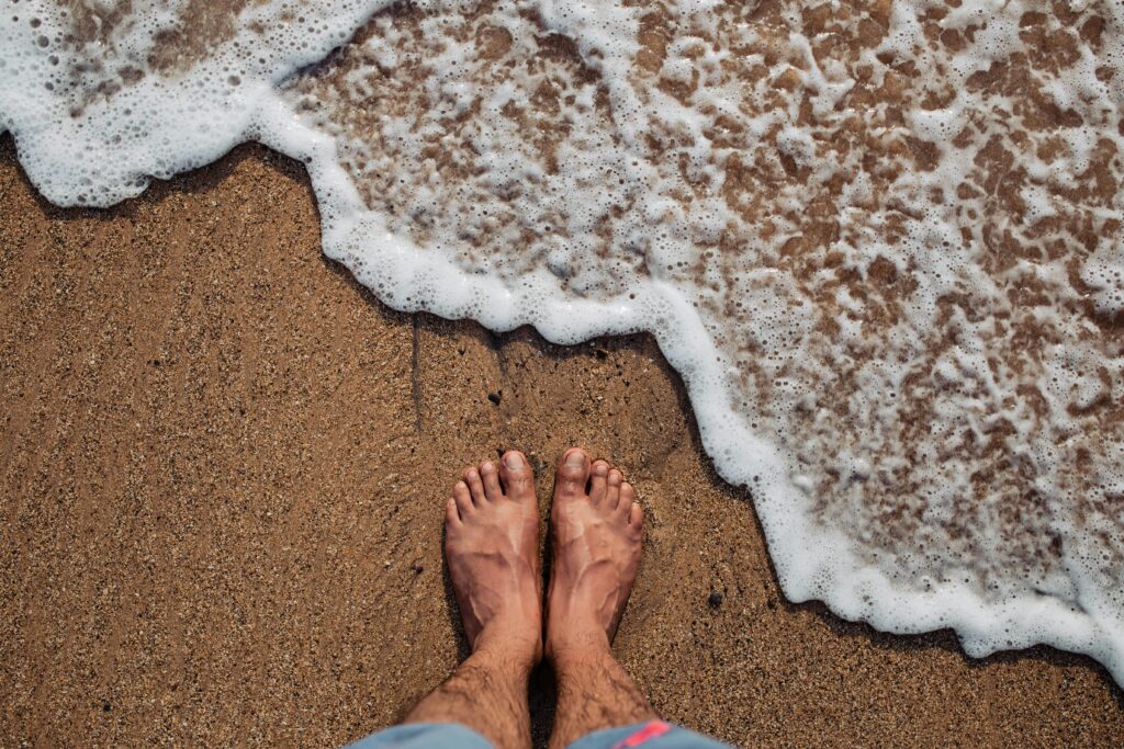 Feet standing on Morjim Beach with sea foam washing ashore. A serene beach moment in India.